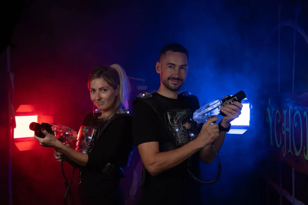 lasergame-karlovy-vary-dva-hraci Dva hráči stojí zády k sobě s laserovými pistolemi v červeno-modře osvětlené aréně.